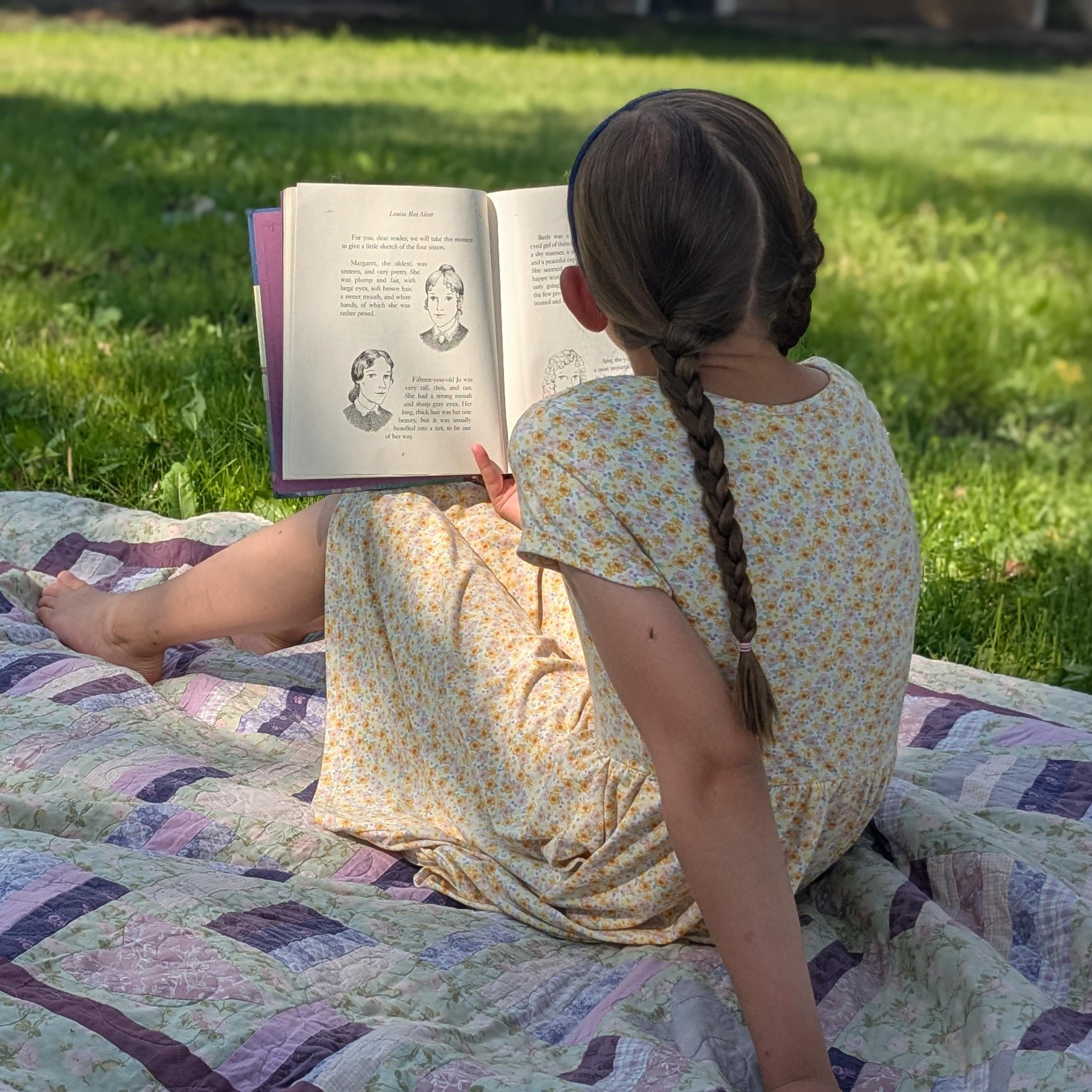 A child reading a book on a picnic blanket in a grassy park