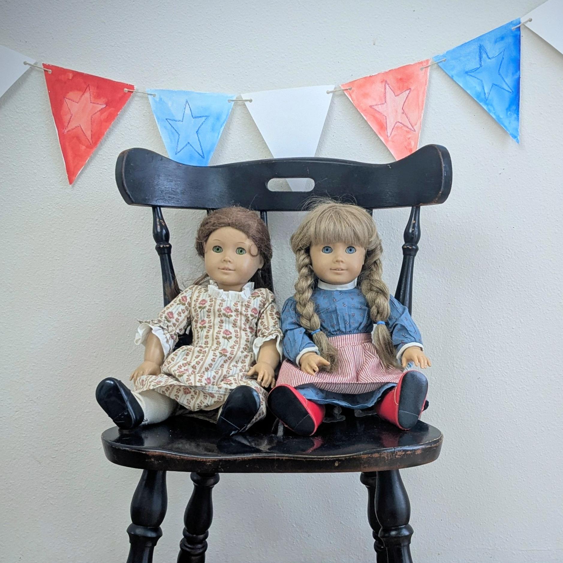 Two dolls sitting on a chair under a red, white, and blue banner